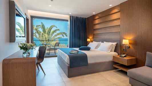 Et hotellrom med seng, nattbord, skrivebord og store glassdører som fører til en balkong med utsikt over palmer og havet. (AI-generert tekst)