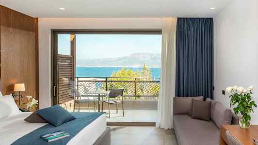 Hotellrom med seng, sofa, skrivebord og utsikt over havet fra en balkong. (AI-generert tekst)