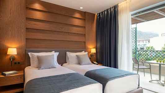 Hotellrom med to enkeltsenger, dekorert med blå sengetepper og grå puter, vegglamper og utsikt mot en balkong. (AI-generert tekst)