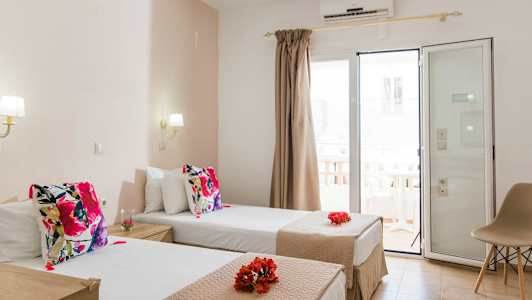 Hotellrom med to enkeltsenger dekorert med fargerike puter og blomster. Glassdør med gardiner som fører til balkong. (AI-generert tekst)