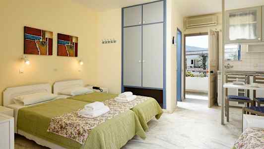 To senger med grøn sengetøy og blomstrete sengeoverkast i et rom med skap, kjøkkenbord og dør til balkong. (AI-generert tekst)