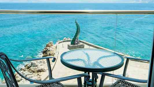 Utsikt fra en terrasse mot sjøen med to stoler og et bord i forgrunnen. (AI-generert tekst)