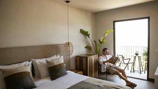 Et hotellrom med en oppredd dobbeltseng, moderne interiør, og en balkong med utemøbler samt grønt. (AI-generert tekst)