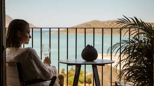En person sitter på en balkong og holder et glass med utsikt over havet. (AI-generert tekst)