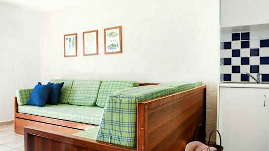 En tresofa med grønne rutemønstrede puter, to blå puter og tre innrammede bilder på veggen. En del av et kjøkken med rutete fliser er synlig. (AI-generert tekst)