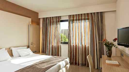 Et hotellrom med en oppredd seng, skrivebord med stol, en TV og et vindu med gardiner. (AI-generert tekst)