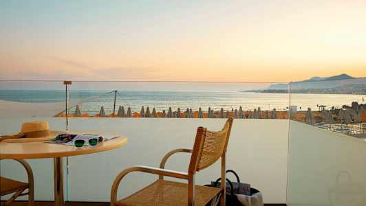 Utsikt fra en terrasse mot havet med solnedgang og strandparasoller i bakgrunnen. (AI-generert tekst)