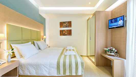 Et hotellrom med en stor seng, dekorert med stripete sengeteppe og hvite puter. På veggen er det kunst med blomstermotiver, og til høyre henger det en TV samt et lite møbel med blomster. (AI-generert tekst)