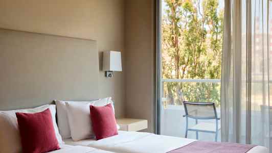 Hotellrom med to senger, røde puter og utsikt mot en balkong med stoler og trær utenfor. (AI-generert tekst)