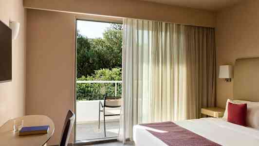 Et hotellrom med en dobbeltseng og en arbeidsplass ved siden av. Rommet har et stort vindu som leder ut til en balkong med en stol og utsikt mot grøntområder. (AI-generert tekst)