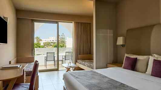 Et moderne hotellrom med en dobbeltseng, skrivebord, sittemøbler og et stort vindu som fører ut til en balkong med utsikt over en hage. (AI-generert tekst)