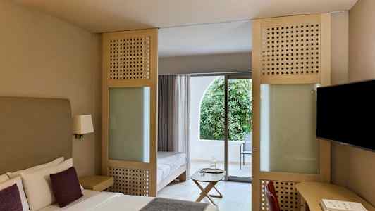 Hotellrom med dobbeltseng, sittemøbler og glassdør som fører til en balkong med utsikt over grønne omgivelser. (AI-generert tekst)