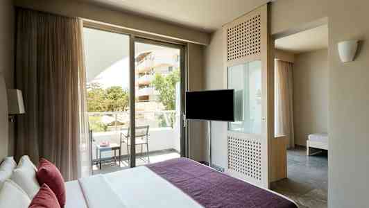 Et hotellrom med en seng, en TV, en balkong med stoler og bord samt en utsikt over grøntområder og et hus. (AI-generert tekst)