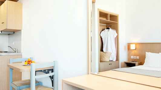 Et lyst hotellrom med en liten kjøkkenkrok, et bord med to stoler og en seng. En hvit kjole henger i skapet. (AI-generert tekst)