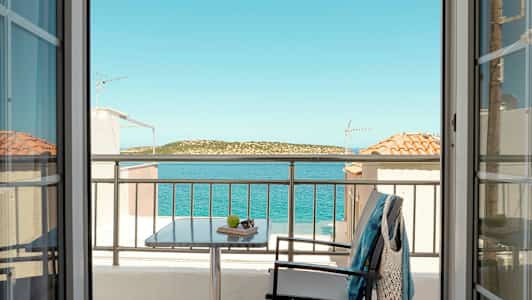 Utsikt fra et rom gjennom dører mot en balkong med bord, stol og klær, samt hav og landskap i bakgrunnen. (AI-generert tekst)