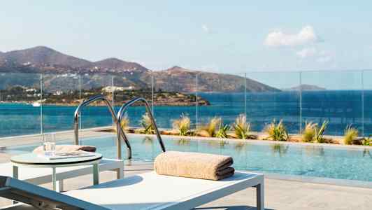 Et utendørs basseng med utsikt over havet og fjellene, omgitt av glassrekkverk og solstoler. (AI-generert tekst)