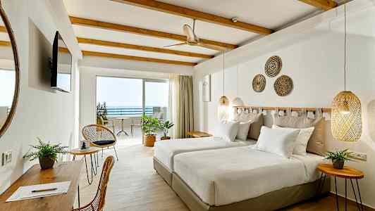 Et hotellrom med to enkeltsenger, veggdekorasjoner og møbler i lyse farger. Utsikt mot en uteplass og havet. (AI-generert tekst)