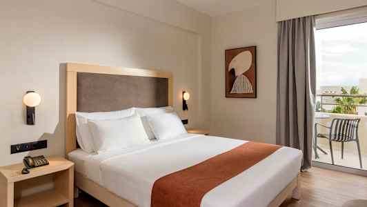 Hotellrom med en dobbeltseng med hvitt sengetøy og brun dekorasjon, et sidebord med telefon, en vegglampe og en uteplass med bord og stol i bakgrunnen. (AI-generert tekst)