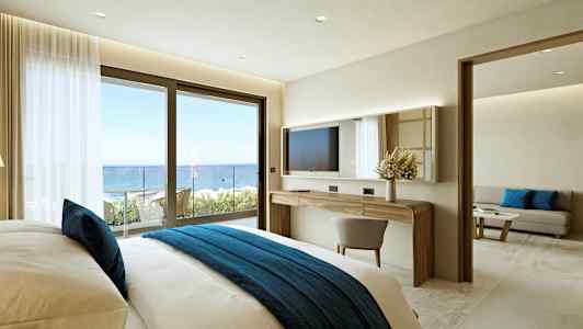 Et hotellrom med en komfortabel seng med blått sengeteppe, et skrivebord med speil, og en skyvedør som leder til en balkong med havutsikt. (AI-generert tekst)