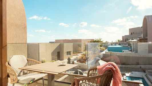 Utsikt fra en terrasse med glassrekkverk. To kurvstoler står ved et lite bord. Utsikt over moderne bygningsstrukturer, et basseng og terrasseområder under en klar blå himmel. (AI-generert tekst)