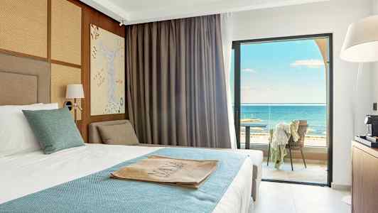 Hotellrom med en oppredd seng med hvitt og blått sengetøy. Rommet har store vinduer med utsikt over havet og en møblert balkong. (AI-generert tekst)