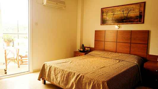 Et soverom med en seng, et nattbord og et maleri på veggen; ved siden av er en åpen balkongdør. (AI-generert tekst)