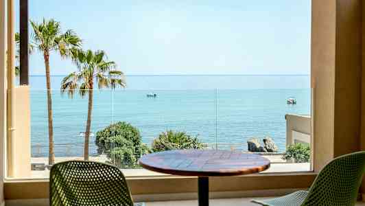 Utsikt fra et rom mot havet, med palmer og stoler på en terrasse. (AI-generert tekst)