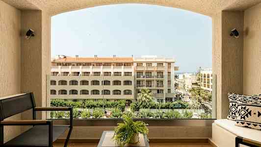 Utsikt fra en balkong på et hotell med sofamøbler, planter og bygninger og trær i bakgrunnen. (AI-generert tekst)