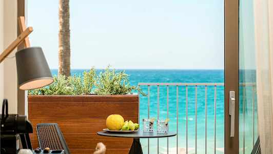 Utsikt fra en balkong mot havet med et bord som har frukt og drikke på seg. (AI-generert tekst)