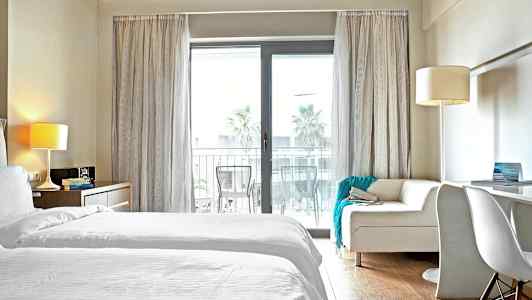 Et hotellrom med hvitt sengetøy og en sofa foran en glassdør til en balkong. (AI-generert tekst)