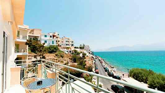 Utsikt fra en balkong mot havet med biler parkert langs en vei og bygninger i nærheten. (AI-generert tekst)