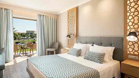 Et hotellrom med en stor seng, dekorative puter og en åpning mot en balkong med utsikt over hagen. (AI-generert tekst)