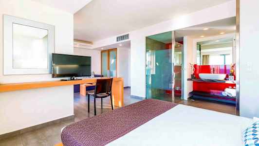 Et moderne hotellrom med en seng i forgrunnen, TV, skrivebord og et åpent baderomsdesign med en rød aksentvegg. (AI-generert tekst)