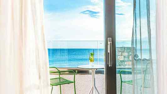 Utsikt fra et rom mot en balkong med bord og stoler, havet i bakgrunnen. (AI-generert tekst)