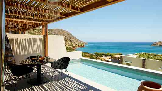 Utsikt over en terrasse med basseng, bord og stoler samt havet og fjellene i bakgrunnen. (AI-generert tekst)