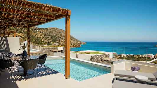 Bilde av en terrasse med basseng, solsenger og spiseplass, med utsikt mot havet og fjell under en klar himmel. (AI-generert tekst)