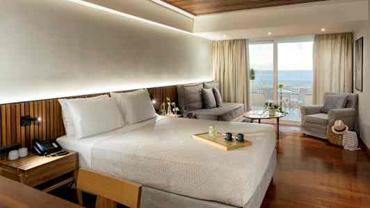 Et hotellrom med en dobbeltseng, en sittegruppe med sofa og lenestol, samt en balkong med utsikt over havet. (AI-generert tekst)