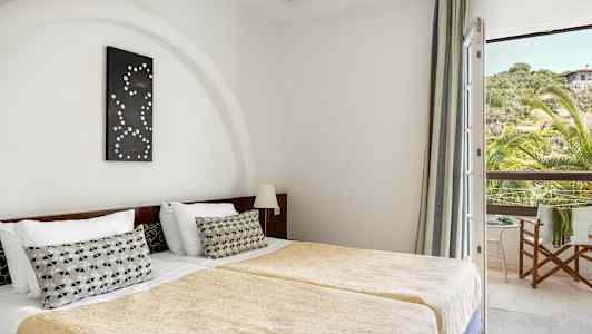 Rommet viser to enkeltsenger med gullfargede sengetepper og en dekorativ pute på hver seng. En dekorasjon henger over hodegavlen, og et vindu med utsikt over grøntområder og et hus ses til høyre. (AI-generert tekst)