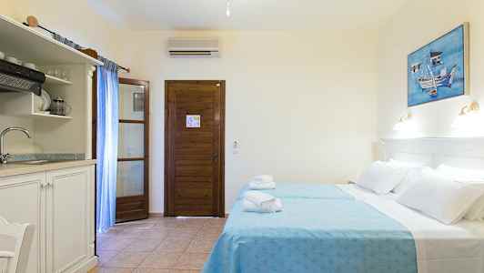 Et hotellrom med to senger kledd i blå sengetøy, en tredør, et lite kjøkken med vask og et maleri på veggen. (AI-generert tekst)