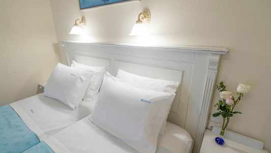 En oppredd dobbeltseng med hvite laken, et blått sengeteppe, hvite puter, et bilde over og en liten vase med blomster på et nattbord. (AI-generert tekst)