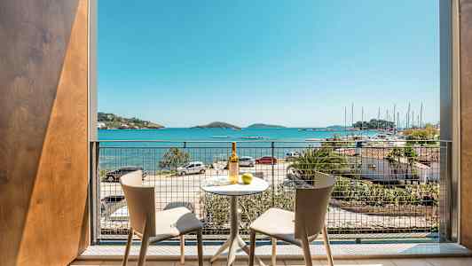 Utsikt fra en balkong med to stoler og et bord, mot havet og en småbåthavn. (AI-generert tekst)