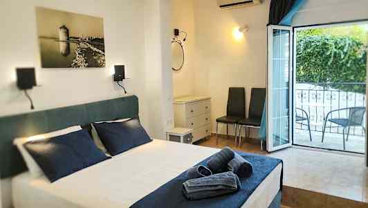 Et hotellrom med en oppredd seng og mørkeblå sengetøy, et åpent vindu mot balkongen, og møbler inkludert to stoler og en kommode. (AI-generert tekst)