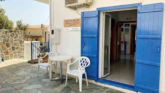 En hvit bygning med blå dører og skodder. Foran døren står to hvite plaststoler og et lite bord. (AI-generert tekst)