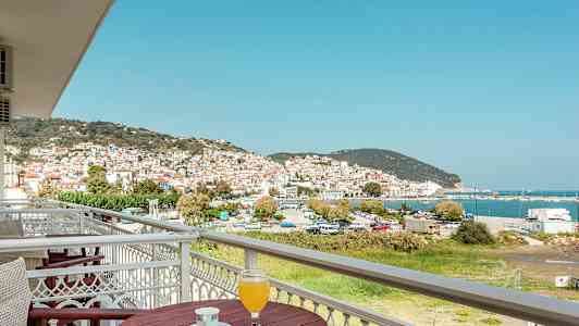 Utsikt over en by med hvite hus og røde tak, ved havet, fra en balkong med bord og drikke. (AI-generert tekst)