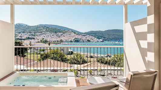 Utsikt fra en terrasse med to stoler og et boblebad, mot en kystby med fjell i bakgrunnen. (AI-generert tekst)