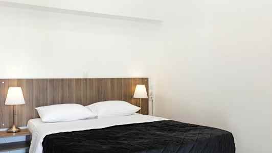 Et soverom med en dobbeltseng, hvitt sengetøy og et mørkt sengeteppe. To sengelamper står på nattbordene ved siden av sengen. (AI-generert tekst)