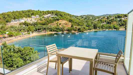 Utsikt fra en terrasse med stoler og bord mot en vik med klart blått vann, omgitt av grønne åser og bygninger. (AI-generert tekst)