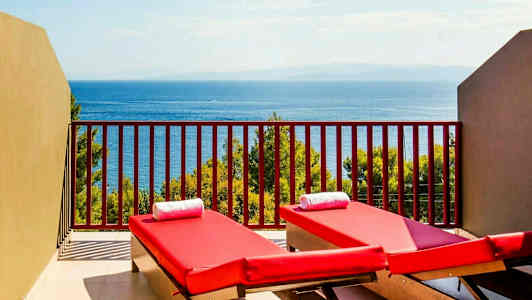 To røde solsenger med hvite håndklær på en balkong med utsikt over havet og vegetasjon. (AI-generert tekst)