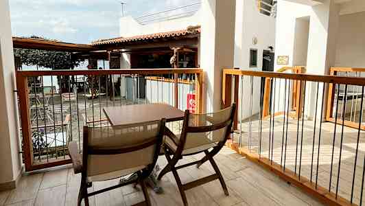 En uteplass med to trestoler og et bord. Utsikt over en bygning og et tak. I bakgrunnen sees en del av havet. (AI-generert tekst)