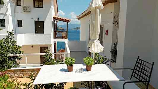 En hvit husbygning med utsikt mot sjøen, et hagebord med to potter og en sammenklappet parasoll. (AI-generert tekst)